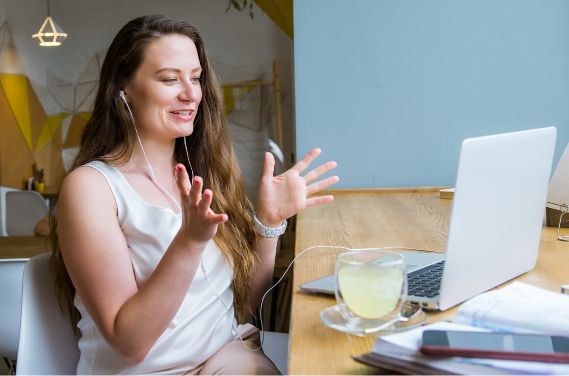 Woman having a video call on a laptop, sitting at a wooden table in a bright room, wearing earphones, with a beverage and documents nearby.