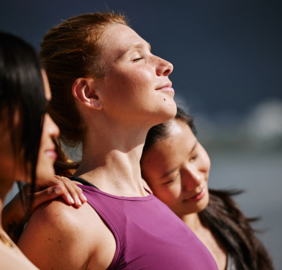 Three individuals stand close together outdoors. The person in the middle, wearing a purple tank top, has eyes closed and face towards the sun. The other two lean on each shoulder.