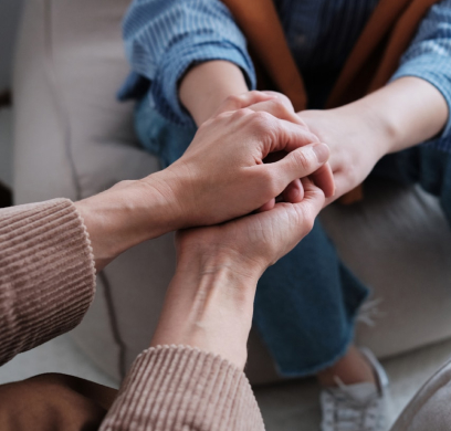 Close-up of two people sitting on a couch, one holding the other's hands. Both wear casual clothing.