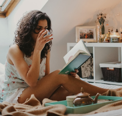 Person drinking from a cup while reading a book, sitting on a bed with a teal tray and a teapot in the foreground.