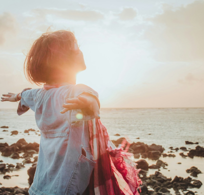 Person with short hair stands on rocky shore, facing the sun with arms outstretched, wearing a denim shirt and holding a scarf. The sun is low, creating a warm light over the ocean.