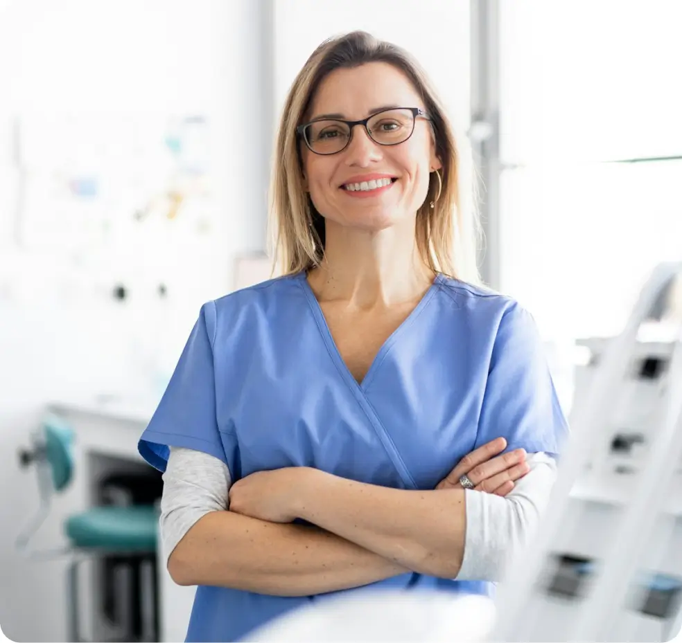 A woman in glasses and blue scrubs stands confidently with arms crossed in a bright room.